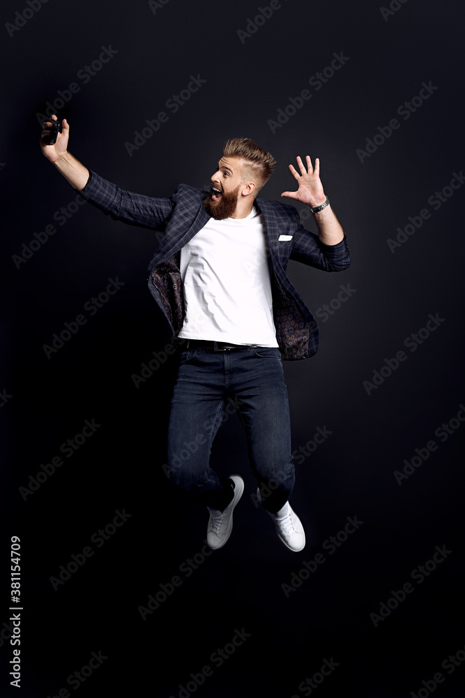 Time to take selfie. Full length of handsome young bearded man in fashion clothes taking selfie while jumping against black background