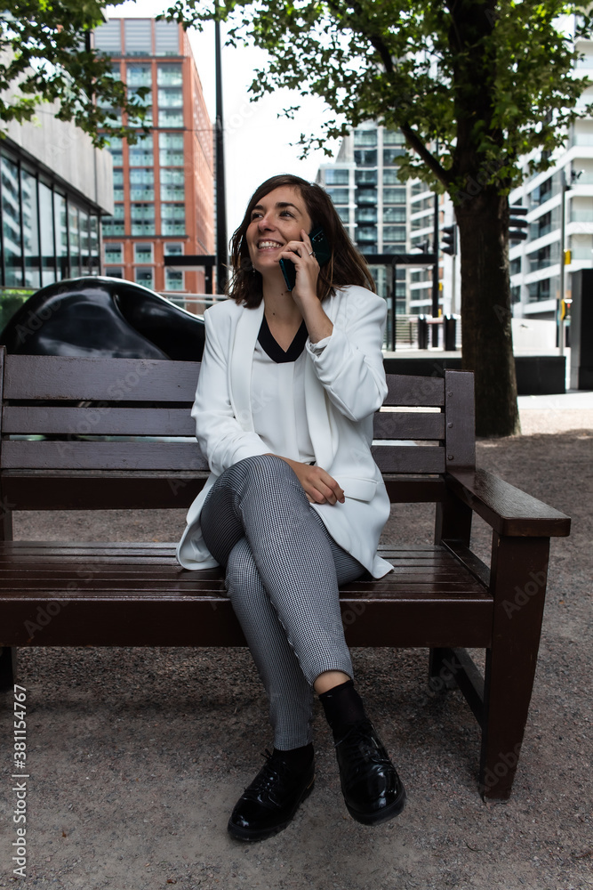 Fototapeta premium Attractive young female wearing a white suit and talking on the phone while sitting on a bench waiting to start a meeting at her office in London, United Kingdom