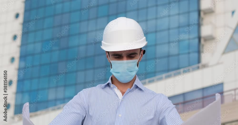 An engineer in a white protective helmet and a face mask holds an unfolded poster with a drawing in front of him, looks at the poster and then to the side against the background of a blue building. An engineer in a white protective helmet and a face mask holds an unfolded poster with a drawing in front of him, looks at the poster and then to the side against the background of a blue building.