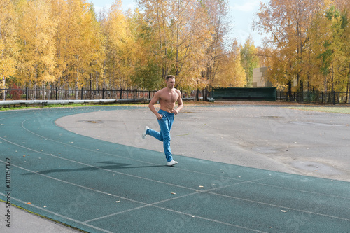 sporty shirtless man jogging on a stadium in fall season. autumn trees on a background. Handsome middle age fit sporty male athlete running to warm up before workout.
