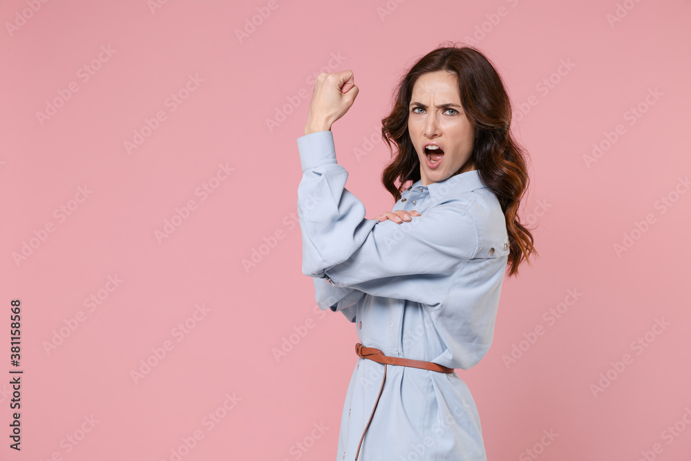 Side view of shocked strong young brunette woman 20s wearing casual blue shirt dress posing standing showing biceps muscles looking camera isolated on pastel pink colour background studio portrait. Side view of shocked strong young brunette woman 20s wearing casual blue shirt dress posing standing showing biceps muscles looking camera isolated on pastel pink colour background studio portrait.