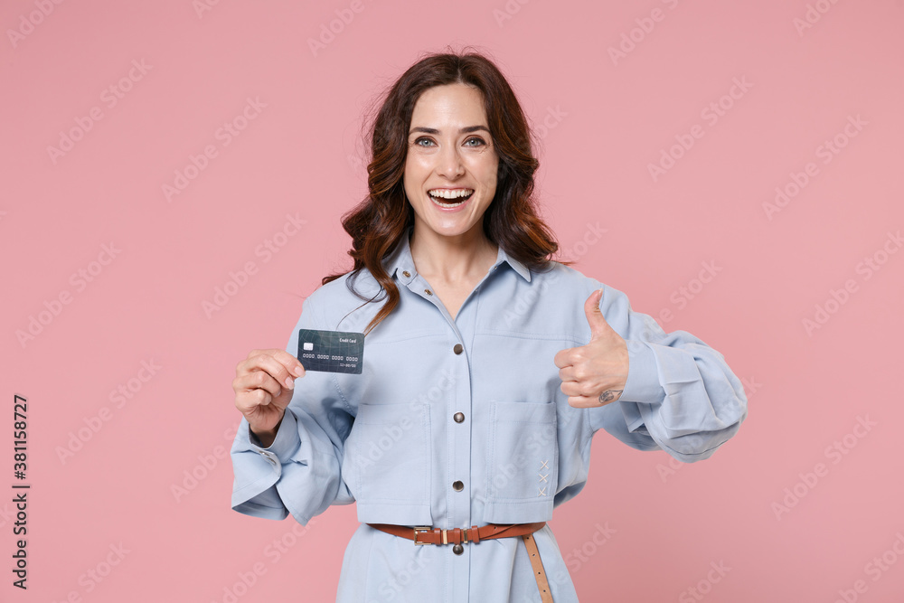 Cheerful young brunette woman 20s wearing casual blue shirt dress posing holding in hand credit bank card showing thumb up looking camera isolated on pastel pink colour background, studio portrait.