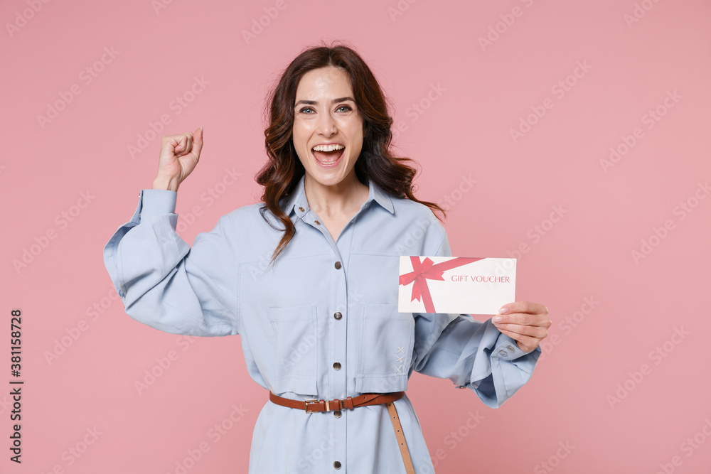 Happy young brunette woman 20s wearing casual blue shirt dress posing holding in hand gift certificate doing winner gesture looking camera isolated on pastel pink colour background, studio portrait. Happy young brunette woman 20s wearing casual blue shirt dress posing holding in hand gift certificate doing winner gesture looking camera isolated on pastel pink colour background, studio portrait.