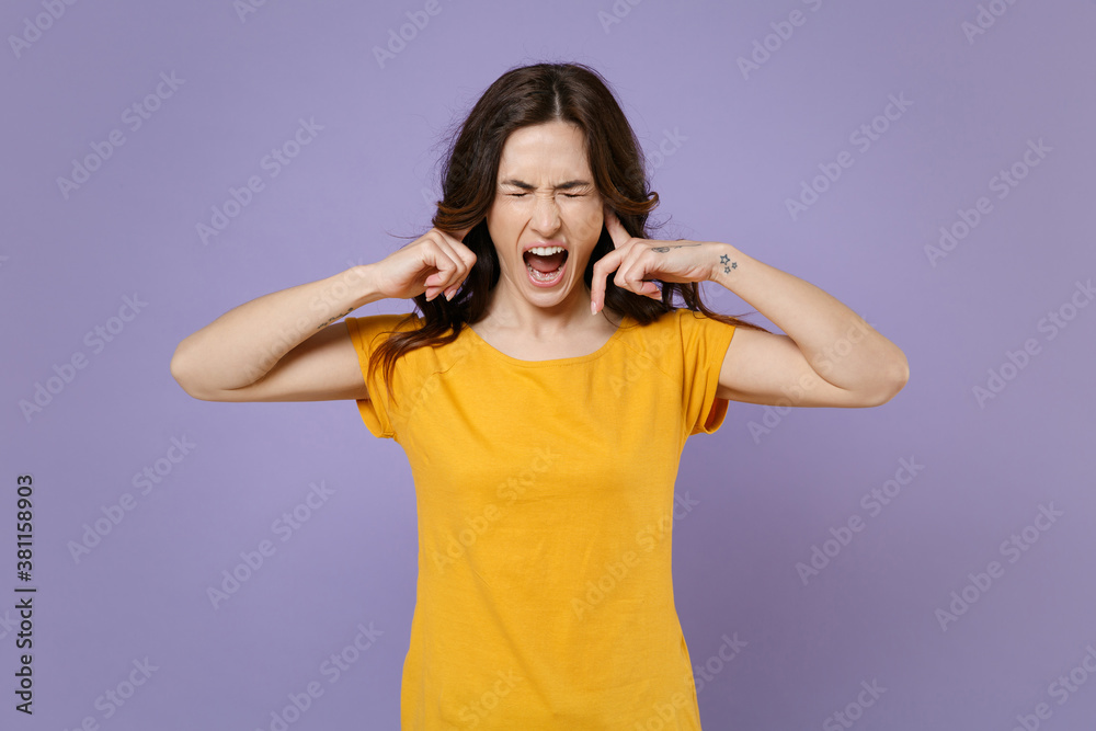 Crazy frustrated young brunette woman 20s wearing basic yellow t-shirt posing covering ears with fingers keeping eyes closed screaming isolated on pastel violet colour background, studio portrait. Crazy frustrated young brunette woman 20s wearing basic yellow t-shirt posing covering ears with fingers keeping eyes closed screaming isolated on pastel violet colour background, studio portrait.