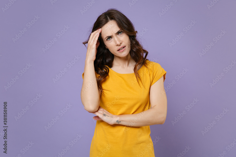 Displeased tired preoccupied puzzled young brunette woman 20s wearing basic yellow t-shirt posing standing put hand on head looking camera isolated on pastel violet colour background, studio portrait. Displeased tired preoccupied puzzled young brunette woman 20s wearing basic yellow t-shirt posing standing put hand on head looking camera isolated on pastel violet colour background, studio portrait.