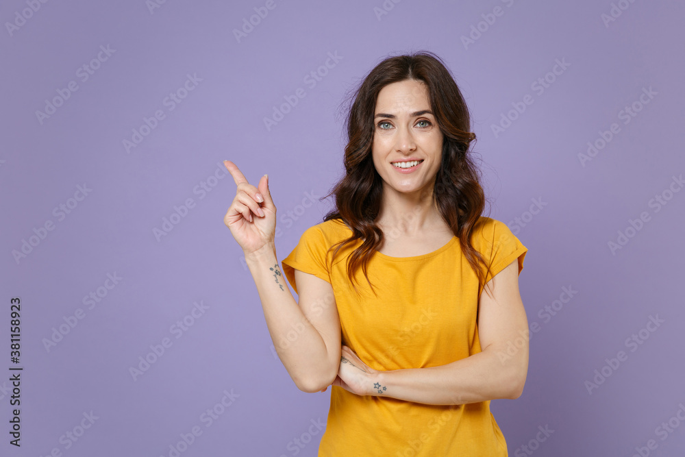 Smiling beautiful young brunette woman 20s wearing basic yellow t-shirt posing stand pointing index finger aside up on mock up copy space isolated on pastel violet colour background, studio portrait. Smiling beautiful young brunette woman 20s wearing basic yellow t-shirt posing stand pointing index finger aside up on mock up copy space isolated on pastel violet colour background, studio portrait.