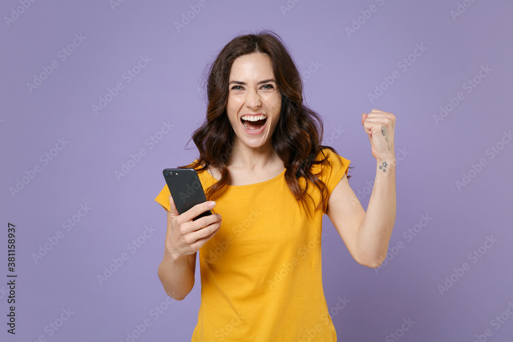 Fototapeta premium Happy young brunette woman 20s wearing basic yellow t-shirt using mobile cell phone typing sms message doing winner gesture looking camera isolated on pastel violet colour background studio portrait.
