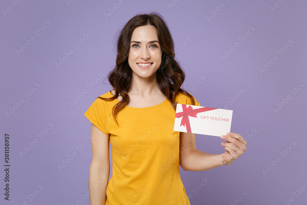 Smiling attractive beautiful young brunette woman 20s wearing basic yellow t-shirt posing hold in hands gift certificate looking camera isolated on pastel violet colour background, studio portrait. Smiling attractive beautiful young brunette woman 20s wearing basic yellow t-shirt posing hold in hands gift certificate looking camera isolated on pastel violet colour background, studio portrait.