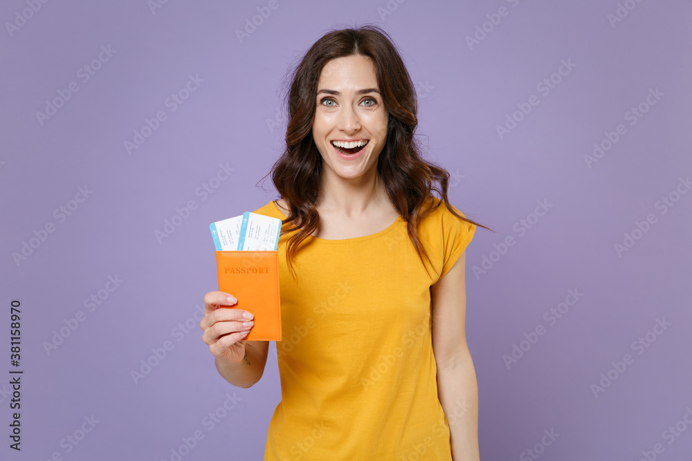 Excited young brunette traveler tourist woman 20s wearing basic yellow t-shirt posing hold passport tickets boarding pass looking camera isolated on pastel violet colour background, studio portrait. Excited young brunette traveler tourist woman 20s wearing basic yellow t-shirt posing hold passport tickets boarding pass looking camera isolated on pastel violet colour background, studio portrait.