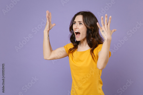 Dissatisfied angry irritated young brunette woman 20s wearing basic yellow t-shirt posing spreading hands screaming swearing looking aside isolated on pastel violet colour background, studio portrait.