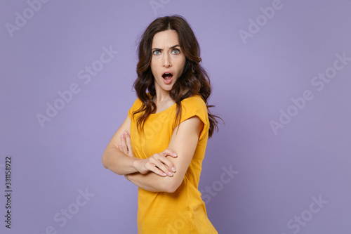 Side view of worried shocked young brunette woman 20s wearing basic yellow t-shirt posing stand holding hands crossed looking camera isolated on pastel violet colour background, studio portrait.
