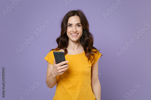 Smiling attractive young brunette woman 20s wearing basic yellow t-shirt posing using mobile cell phone typing sms message looking camera isolated on pastel violet colour background, studio portrait.