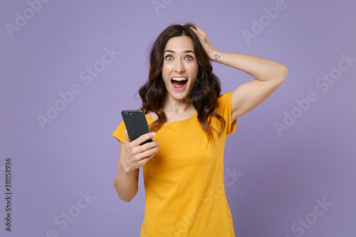 Shocked young brunette woman 20s wearing basic yellow t-shirt using mobile cell phone typing sms message put hand on head looking camera isolated on pastel violet colour background studio portrait.