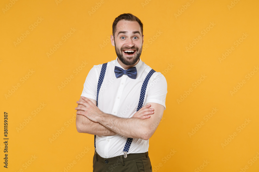 Excited young bearded man 20s wearing white shirt bow-tie suspender posing holding hands crossed keeping mouth open looking camera isolated on bright yellow color wall background, studio portrait. Excited young bearded man 20s wearing white shirt bow-tie suspender posing holding hands crossed keeping mouth open looking camera isolated on bright yellow color wall background, studio portrait.