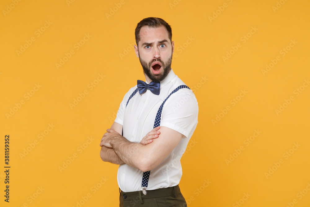 Side view of shocked perplexed young bearded man 20s wearing white shirt bow-tie suspender posing standing holding hands crossed looking camera isolated on yellow color background studio portrait. Side view of shocked perplexed young bearded man 20s wearing white shirt bow-tie suspender posing standing holding hands crossed looking camera isolated on yellow color background studio portrait.
