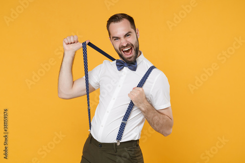 Cheerful funny screaming young bearded man 20s wearing white shirt bow-tie posing standing stretching suspender looking camera isolated on bright yellow color wall background studio portrait.