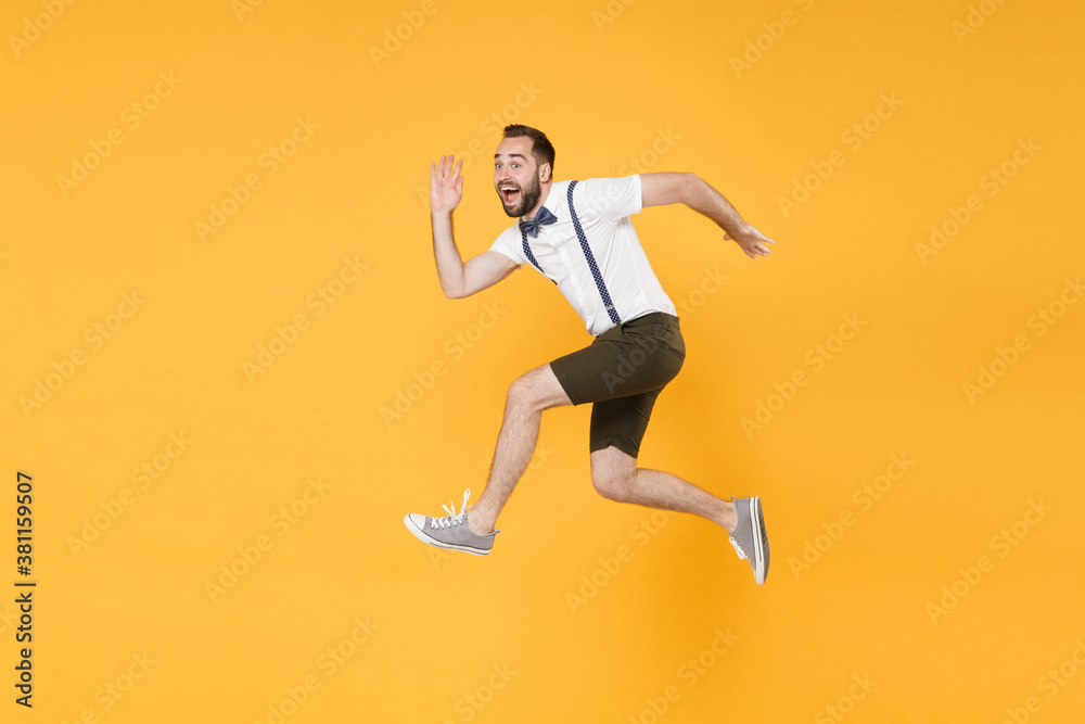Full length side view portrait of cheerful young bearded man 20s wearing white shirt suspender shorts posing jumping like running looking camera isolated on bright yellow color wall background studio.