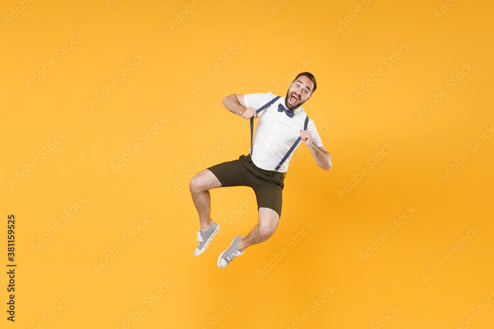 Full length portrait of excited cheerful young bearded man 20s wearing white shirt shorts posing jumping stretching suspender looking camera isolated on bright yellow color wall background studio.