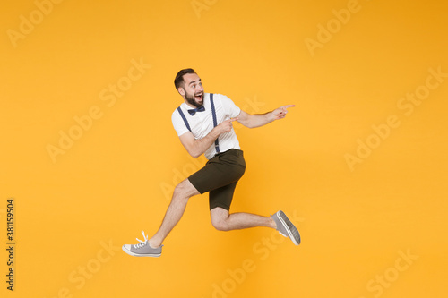 Full length side view portrait of excited young bearded man 20s in white shirt suspender shorts posing jumping pointing index fingers aside on mock up copy space isolated on yellow background studio.