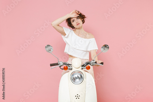 Exhausted tired dissatisfied young woman 20s wearing white summer clothes posing put hand on head looking camera sitting driving moped isolated on pastel pink colour background studio portrait. photo