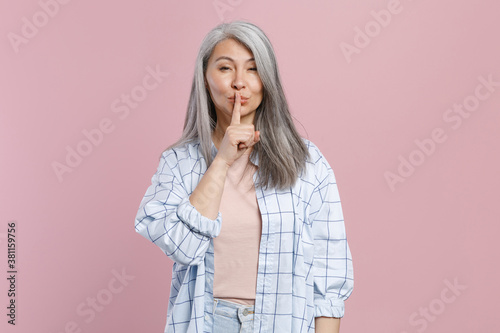 Secret gray-haired asian woman wearing basic white checkered shirt stand saying hush be quiet with finger on lips shhh gesture looking camera isolated on pastel pink colour background studio portrait.