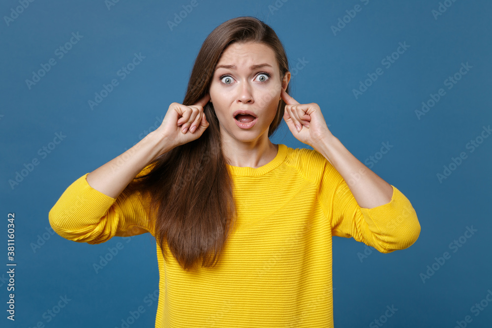 Shocked worried dissatisfied young brunette woman 20s in yellow casual clothes posing covering ears with fingers keeping mouth open looking camera isolated on blue color background studio portrait. Shocked worried dissatisfied young brunette woman 20s in yellow casual clothes posing covering ears with fingers keeping mouth open looking camera isolated on blue color background studio portrait.