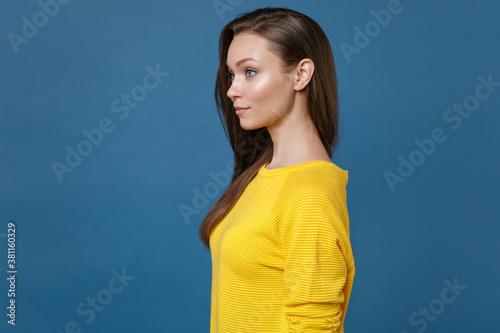 Side view of smiling beautiful attractive charming young brunette woman 20s wearing yellow casual clothes posing standing and looking aside isolated on blue color wall background studio portrait.