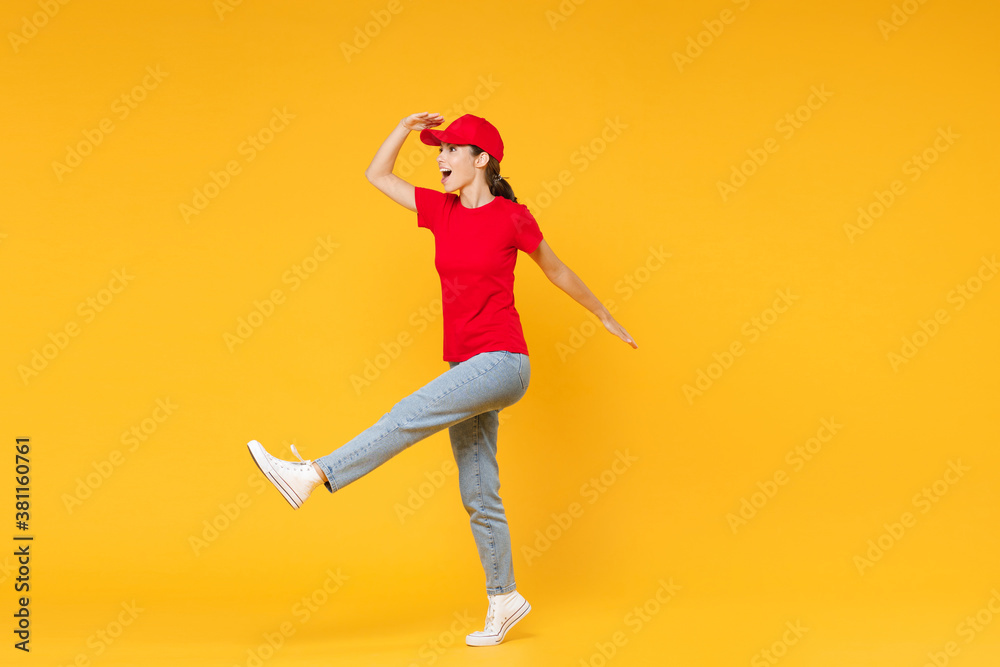 Full length body delivery employee woman in red cap blank t-shirt uniform work courier in service during quarantine coronavirus covid-19 virus standing isolated on yellow background studio portrait.