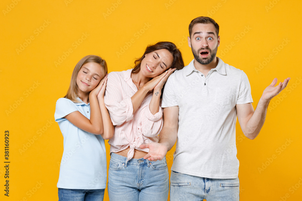 Pretty shocked young parents mom dad with child kid daughter teen girl in t-shirts sleep with folded palms under cheek spreading hand isolated on yellow background studio portrait. Family day concept. Pretty shocked young parents mom dad with child kid daughter teen girl in t-shirts sleep with folded palms under cheek spreading hand isolated on yellow background studio portrait. Family day concept.