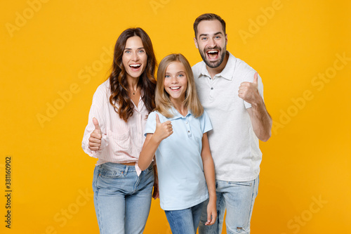 Cheerful funny young happy parents mom dad with child kid daughter teen girl in basic t-shirts showing thumbs up isolated on yellow background studio portrait. Family day parenthood childhood concept.