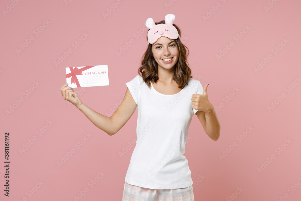Smiling young woman 20s in pajamas home wear sleep mask showing thumb up holding gift certificate while resting at home isolated on pink background studio portrait. Relax good mood lifestyle concept. Smiling young woman 20s in pajamas home wear sleep mask showing thumb up holding gift certificate while resting at home isolated on pink background studio portrait. Relax good mood lifestyle concept.