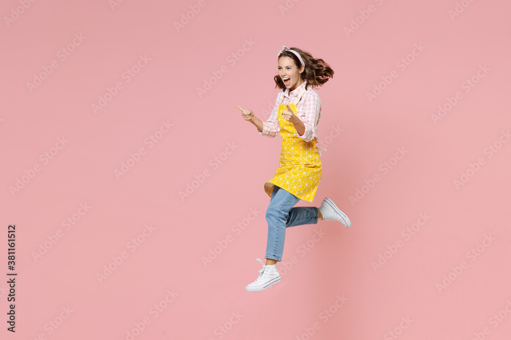 Full length portrait of excited young woman housewife 20s in yellow apron jumping pointing index fingers aside doing housework isolated on pastel pink colour background studio. Housekeeping concept. Full length portrait of excited young woman housewife 20s in yellow apron jumping pointing index fingers aside doing housework isolated on pastel pink colour background studio. Housekeeping concept.