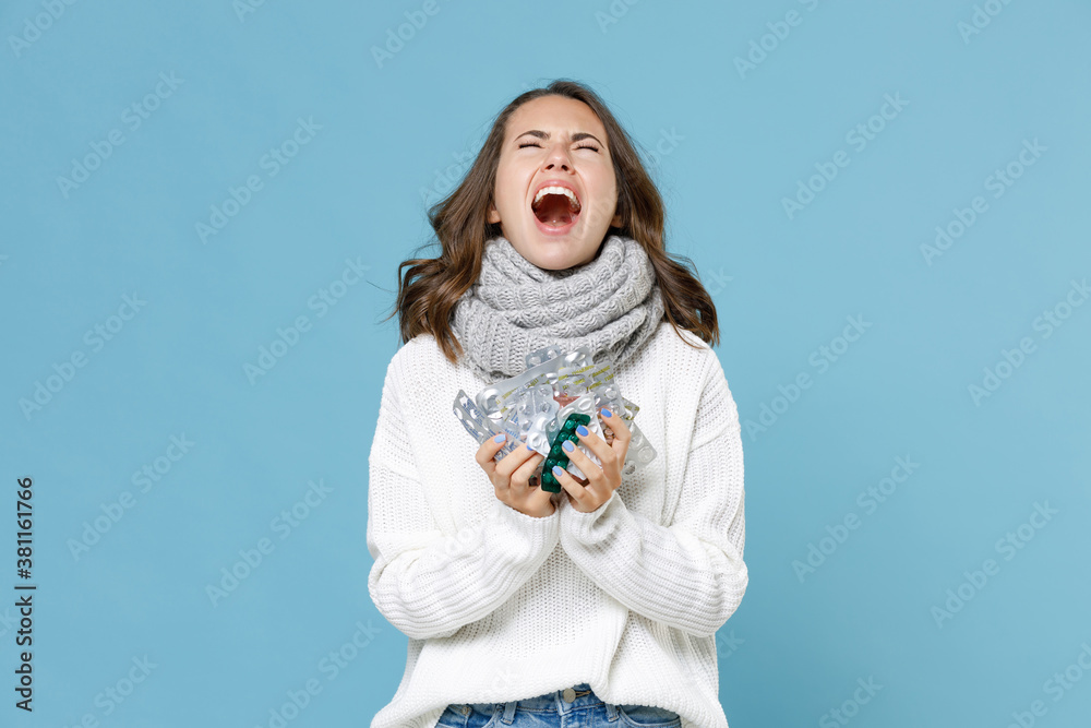Crying screaming young woman wearing white sweater gray scarf hold medication tablets pills isolated on blue background studio portrait. Healthy lifestyle sick disease treatment cold season concept. Crying screaming young woman wearing white sweater gray scarf hold medication tablets pills isolated on blue background studio portrait. Healthy lifestyle sick disease treatment cold season concept.