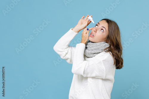 Young brunette woman 20s wearing white sweater gray scarf standing dripping eye drops isolated on blue background studio portrait. Healthy lifestyle ill sick disease treatment cold season concept.