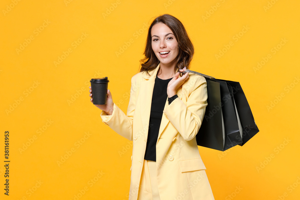 Charming young woman in basic suit jacket hold package bags with purchases after shopping paper cup of coffee or tea looking camera isolated on yellow background studio portrait. Black friday sale. Charming young woman in basic suit jacket hold package bags with purchases after shopping paper cup of coffee or tea looking camera isolated on yellow background studio portrait. Black friday sale.
