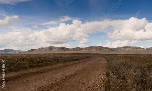 White clouds in the blue sky above the steppe in Khakassia. Wide dirt road going through the steppe up to the hills. 