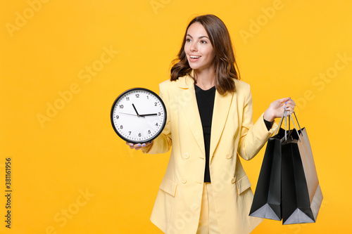 Smiling young brunette woman 20s wearing basic light suit jacket hold package bags with purchases after shopping clock looking aside isolated on yellow background, studio portrait. Black friday sale.