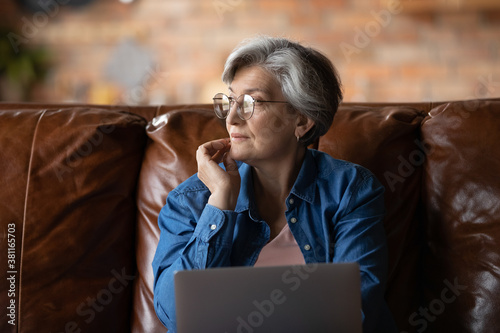 Fotografie Dreamy elderly Caucasian grey-haired woman in glasses distracted from computer w