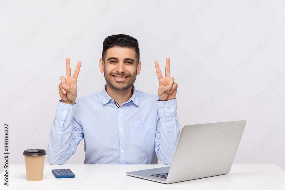 This is win! Joyful businessman sitting office workplace with laptop on desk, showing victory gesture, celebrating successful project, job promotion. indoor studio shot isolated on white background This is win! Joyful businessman sitting office workplace with laptop on desk, showing victory gesture, celebrating successful project, job promotion. indoor studio shot isolated on white background