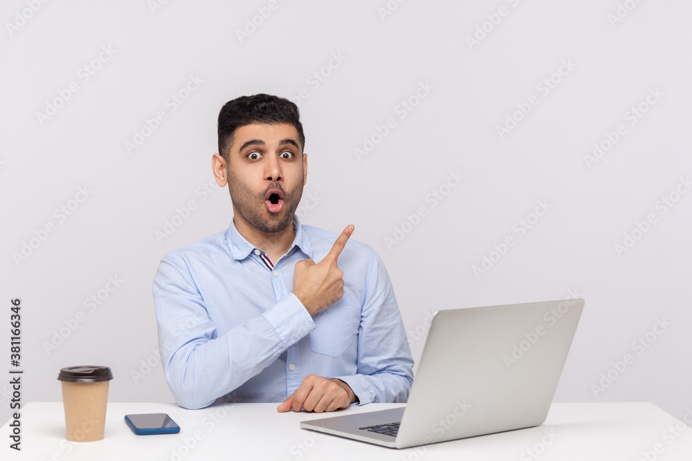 Wow look, advertise here! Amazed shocked businessman sitting office workplace with laptop on desk, pointing aside blank copy space for commercial idea. indoor studio shot isolated on white background Wow look, advertise here! Amazed shocked businessman sitting office workplace with laptop on desk, pointing aside blank copy space for commercial idea. indoor studio shot isolated on white background