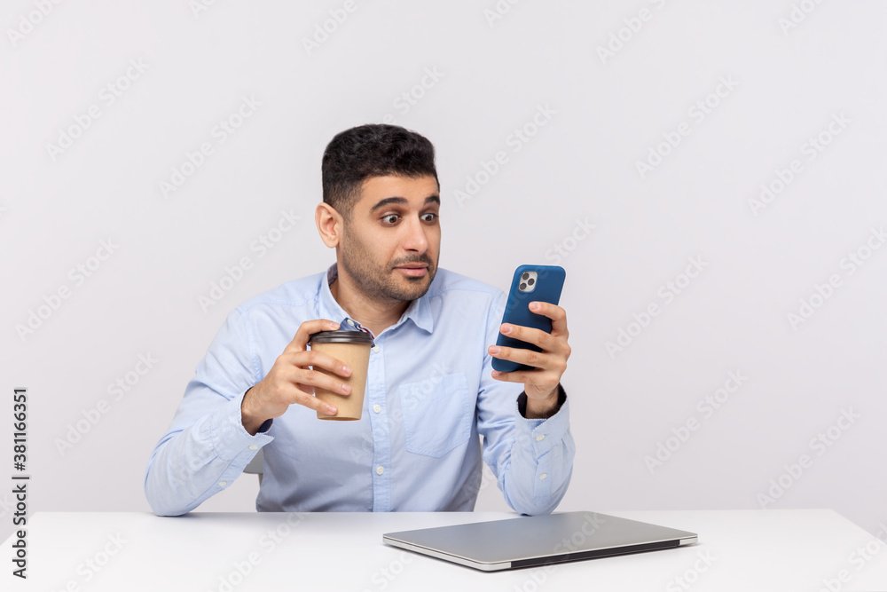 Amazed shocked businessman sitting at workplace with coffee in hand and checking message, reading email on mobile phone with surprised expression. indoor studio shot isolated on white background Amazed shocked businessman sitting at workplace with coffee in hand and checking message, reading email on mobile phone with surprised expression. indoor studio shot isolated on white background
