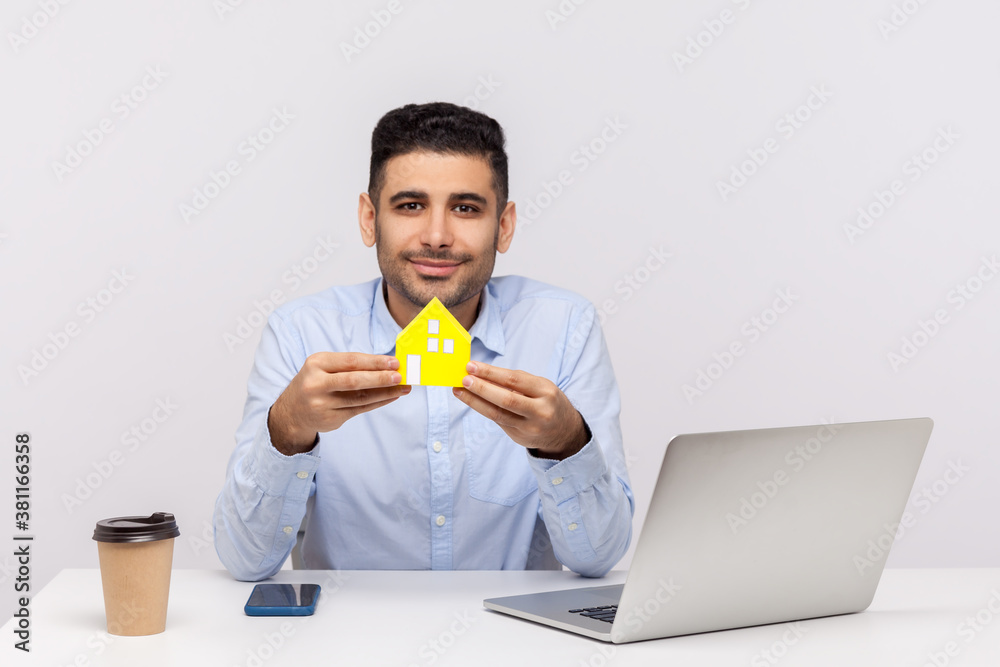 Portrait of real estate agent man sitting in office workplace and holding paper house, smiling friendly, advertisement of purchase or rental housing. indoor studio shot isolated on white background Portrait of real estate agent man sitting in office workplace and holding paper house, smiling friendly, advertisement of purchase or rental housing. indoor studio shot isolated on white background