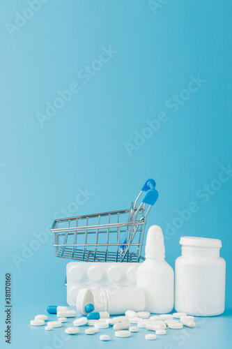 scattered variety pills, drugs, spay, bottles, thermometer, syringe and empty shopping trolley cart on blue background. pharmacy shopping concept