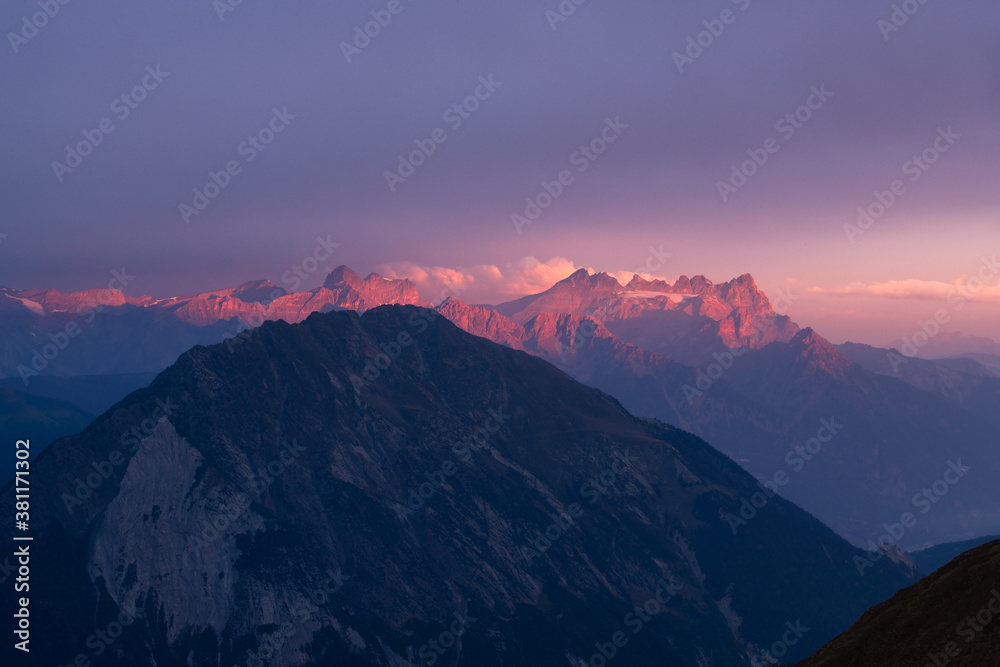 Golden hour scene of warm glowing mountain ridge topped with few clouds partly covered by dark mountain silouhette near Martigny in clear purple sky Golden hour scene of warm glowing mountain ridge topped with few clouds partly covered by dark mountain silouhette near Martigny in clear purple sky