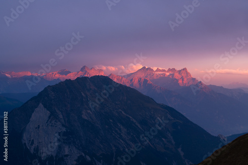 Golden hour scene of warm glowing mountain ridge topped with few clouds partly covered by dark mountain silouhette near Martigny in clear purple sky