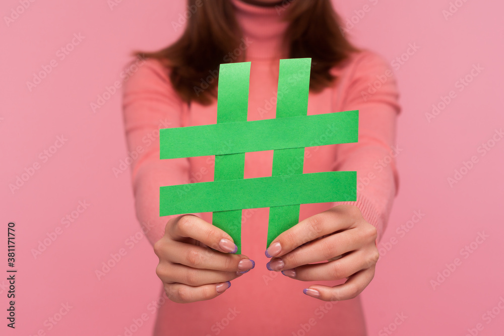 Closeup brunette woman blogger in pink sweater showing green hashtag symbol to camera recommending viral social media topic. Indoor studio shot isolated on pink background Closeup brunette woman blogger in pink sweater showing green hashtag symbol to camera recommending viral social media topic. Indoor studio shot isolated on pink background
