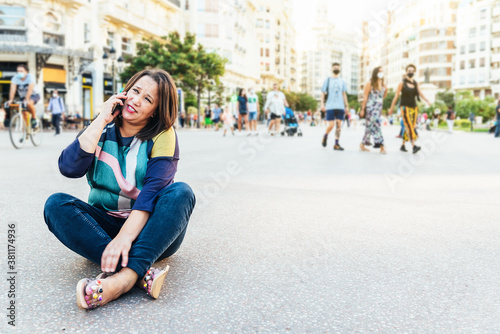 brunette woman in casual clothes sitting on the ground in a square in Europe with people, talking on mobile phone
