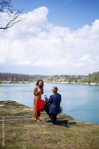 Lake in the forest: a man made a surprise to his girlfriend, he puts a ring on her finger, a declaration of love and an offer of marriage. Girl with a smile on her face and a bouquet of red roses