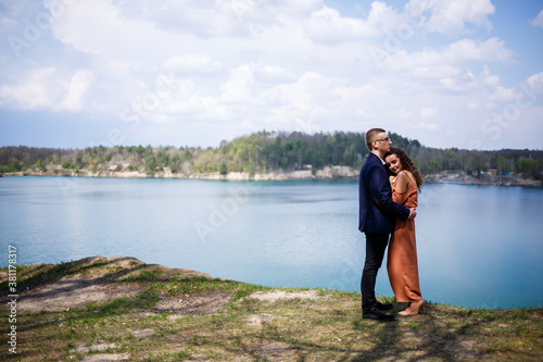 Happy and young newlyweds go holding hands and laughing, on the background of a lake and a green meadow. Groom and beautiful bride with curly hair are walking in the meadow. Joyful summer day © Дмитрий Ткачук
