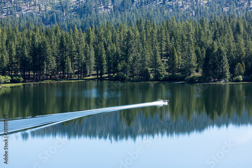 Boat leaves a trail of wake across beautiful blue water with tree and sky reflections